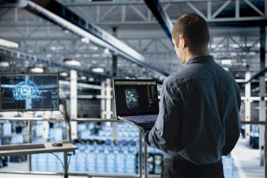 Mitarbeiter steht mit Laptop vor Produktionshalle, auf dem Laptop sieht man Planungsmechanismen